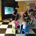 Čas u vrtiću: deca na šarenim stolicama rade na tabletima, roditelji sede iza njih, na zidu je projekcija zadatka.