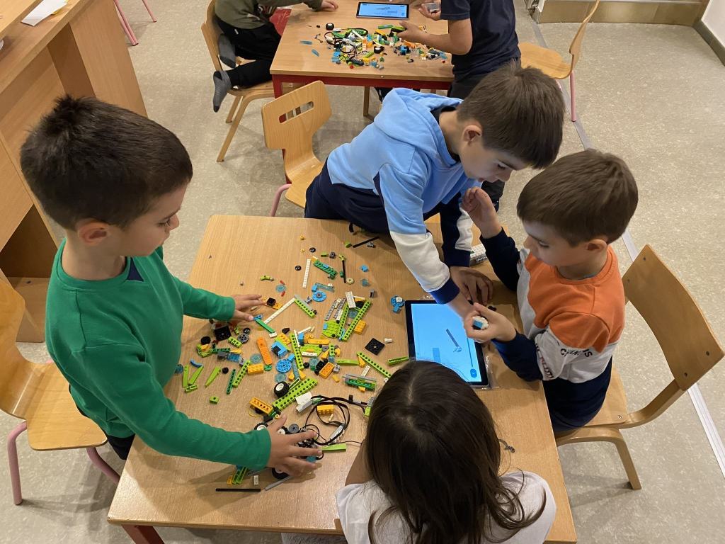 Četvoro dece za stolom sklapa LEGO delove, pored njih stoji tablet